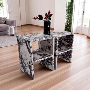 May include: Two matching side tables constructed from white and dark purple veined marble, each featuring a rectangular frame with a single horizontal shelf. A black marble vase containing red berries sits on the surface of the left table.