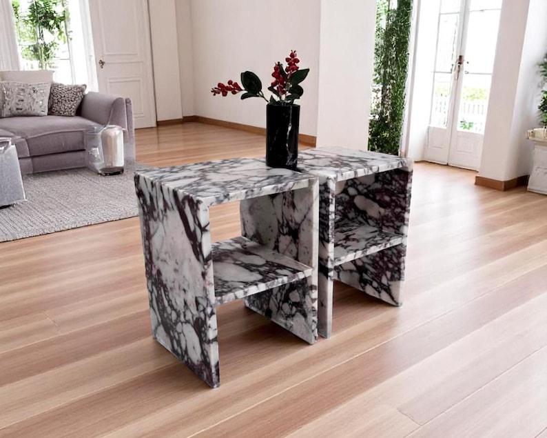 May include: Two matching side tables constructed from white and dark purple veined marble, each featuring a rectangular frame with a single horizontal shelf. A black marble vase containing red berries sits on the surface of the left table.