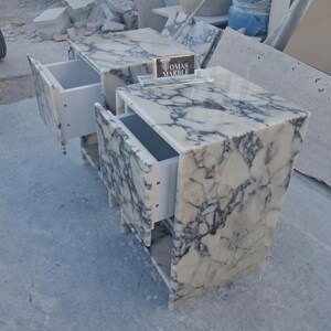 Puede incluir: Dos mesitas de noche de m&aacute;rmol con cajones abiertos. Las mesitas son blancas con vetas grises. Los cajones son blancos. El texto "TOMAS MARBLE" es visible en un peque&ntilde;o letrero entre las mesitas.