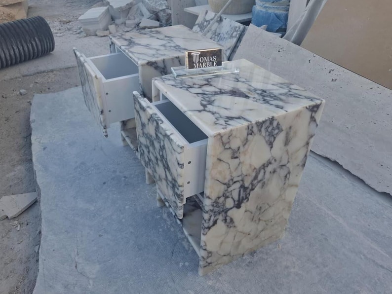 Puede incluir: Dos mesitas de noche de m&aacute;rmol con cajones abiertos. Las mesitas son blancas con vetas grises. Los cajones son blancos. El texto "TOMAS MARBLE" es visible en un peque&ntilde;o letrero entre las mesitas.