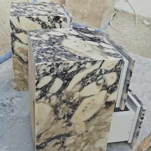 May include: Two rectangular pedestals featuring a polished marble surface with a high-contrast pattern of cream, white, and dark charcoal-black veining. Each unit has a smooth, block-like structure with a visible white interior drawer component.