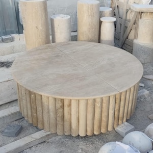 Op de afbeelding: Een ronde, beige stenen tafel met een uniek ontwerp. Het tafelblad is gemaakt van glad, plat steen en de basis is gemaakt van gestapelde, ronde stenen kolommen.