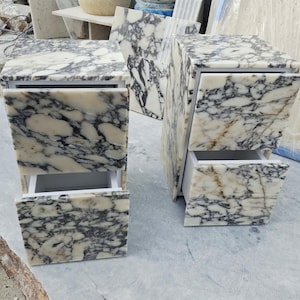 May include: Two rectangular bedside tables crafted from white marble with prominent black and grey veining. Each table features two drawers with matching marble fronts and white interior frames, showcasing a polished stone finish throughout.