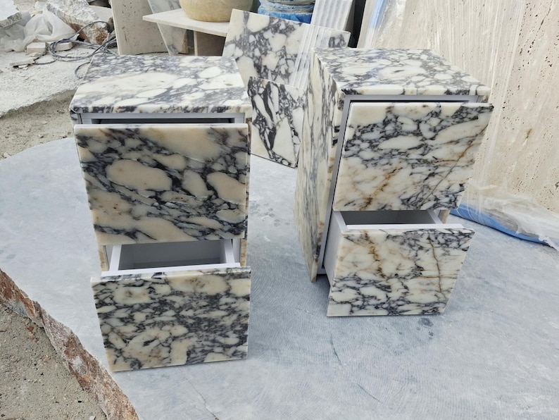 May include: Two rectangular bedside tables crafted from white marble with prominent black and grey veining. Each table features two drawers with matching marble fronts and white interior frames, showcasing a polished stone finish throughout.