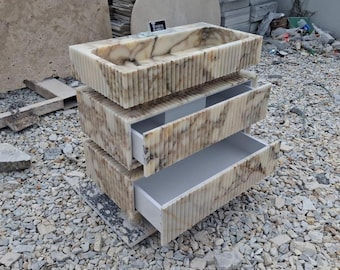 Op de afbeelding: Een rechthoekig wastafelmeubel van beige en bruin geaderd marmer, voorzien van een opzetwastafel en twee open lades. De buitenzijden zijn afgewerkt met een verticale gecanneleerde textuur en een klein zwart label met de tekst Osman Marble.