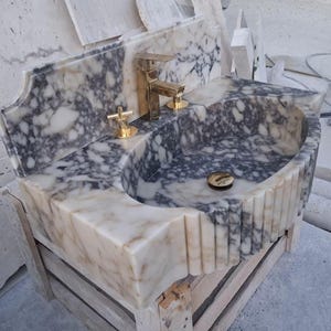 May include: A marble bathroom sink with a gold faucet and fixtures. The sink basin is oval-shaped with a gray and white veined pattern. The sink is set on a wooden base and has a decorative backsplash.
