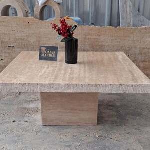 May include: A square travertine coffee table with a beveled edge and a simple, modern design. The table is made of natural stone and has a light beige color. The table is sitting on a concrete floor. The text "Thomas Marble" is visible on a small card on the table.