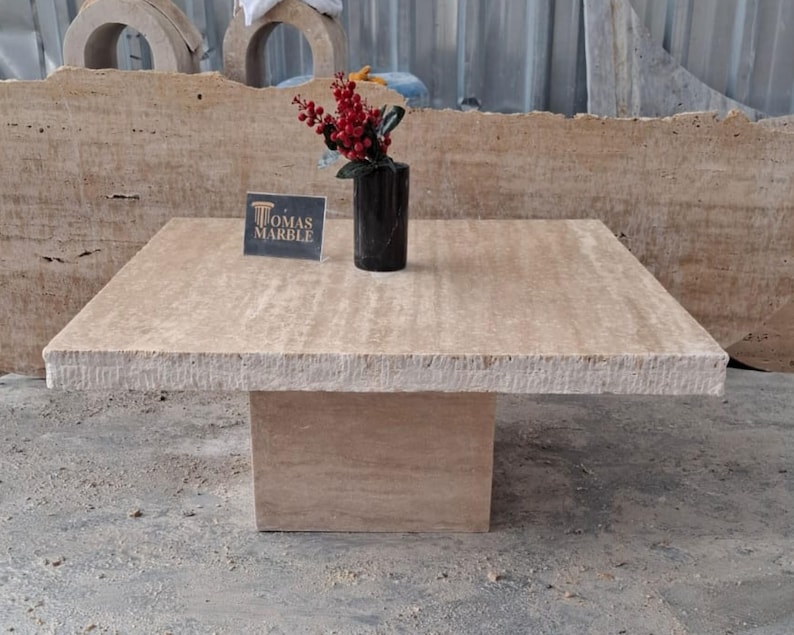 May include: A square travertine coffee table with a beveled edge and a simple, modern design. The table is made of natural stone and has a light beige color. The table is sitting on a concrete floor. The text "Thomas Marble" is visible on a small card on the table.