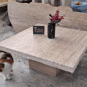 May include: A square, light beige travertine coffee table with a rough, textured surface. The table has a single, rectangular base and is sitting on a concrete floor. A small, black vase with red berries is on the table, and a business card for "Tomas Marble" is also on the table. A cat is sitting on the floor in the foreground.