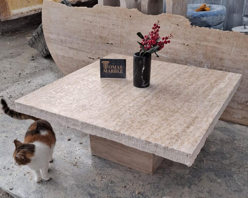 May include: A square, light beige travertine coffee table with a rough, textured surface. The table has a single, rectangular base and is sitting on a concrete floor. A small, black vase with red berries is on the table, and a business card for "Tomas Marble" is also on the table. A cat is sitting on the floor in the foreground.
