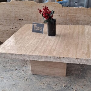 May include: A square, light beige travertine coffee table with a rough, textured edge. The table has a square base and is sitting on a concrete floor. A small vase with red berries is on the table, and a sign that says "Tomas Marble" is in front of the vase.