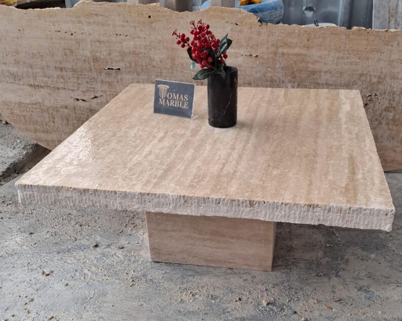 May include: A square, light beige travertine coffee table with a rough, textured edge. The table has a square base and is sitting on a concrete floor. A small vase with red berries is on the table, and a sign that says "Tomas Marble" is in front of the vase.