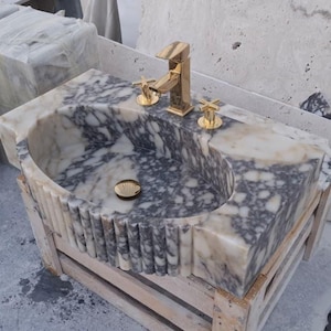 May include: A marble bathroom sink with a gold tap and fixtures. The sink features a grey and white marbled pattern and is set on a wooden crate. The tap and fixtures are gold-coloured.