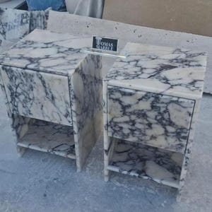 Puede incluir: Dos mesitas de noche de m&aacute;rmol con un patr&oacute;n veteado blanco y gris. Cada mesita de noche tiene un caj&oacute;n y una balda abierta. Las mesitas de noche son de m&aacute;rmol y tienen forma rectangular. Las mesitas de noche est&aacute;n una al lado de la otra.