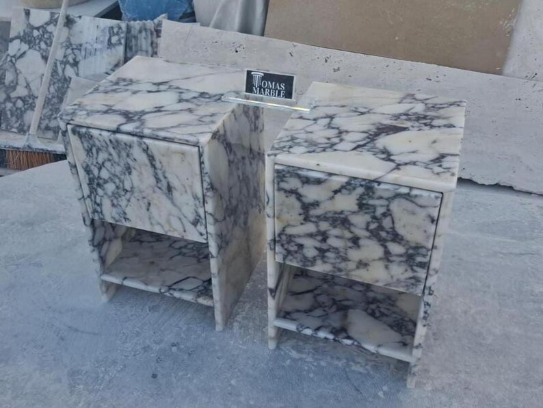 Puede incluir: Dos mesitas de noche de m&aacute;rmol con un patr&oacute;n veteado blanco y gris. Cada mesita de noche tiene un caj&oacute;n y una balda abierta. Las mesitas de noche son de m&aacute;rmol y tienen forma rectangular. Las mesitas de noche est&aacute;n una al lado de la otra.