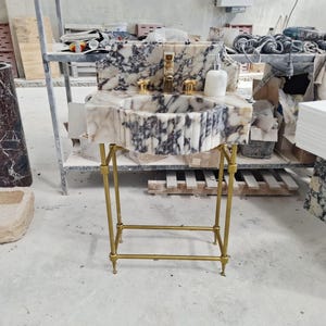 May include: A white and dark gray marble pedestal sink featuring a fluted basin, a high backsplash, and a gold-toned metal stand. The sink includes a gold-toned faucet, two gold-toned handles, and a white marble soap dispenser on the countertop.