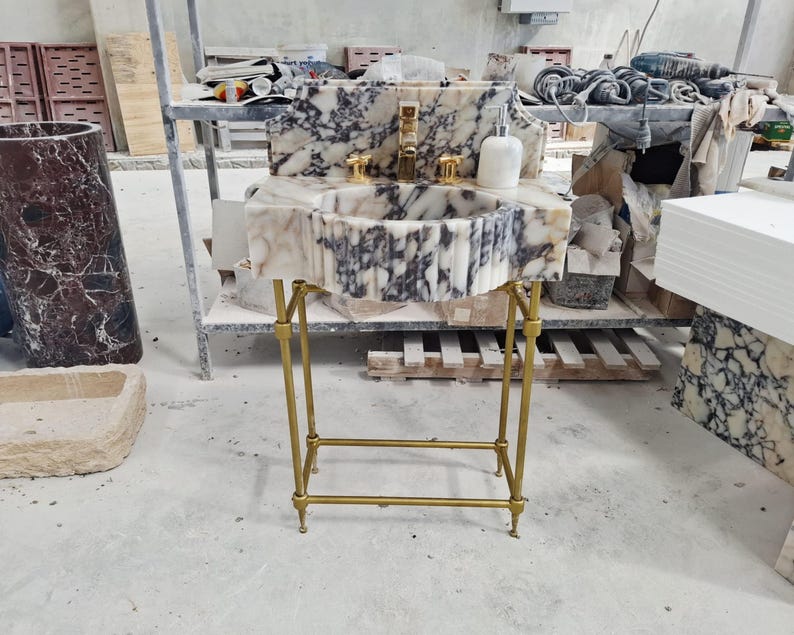 May include: A white and dark gray marble pedestal sink featuring a fluted basin, a high backsplash, and a gold-toned metal stand. The sink includes a gold-toned faucet, two gold-toned handles, and a white marble soap dispenser on the countertop.