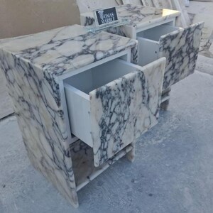Puede incluir: Dos mesitas de noche de m&aacute;rmol con cajones abiertos. Las mesitas son blancas con vetas grises. Los cajones son blancos. El texto "OMAS MARBLE" es visible en un peque&ntilde;o letrero encima.