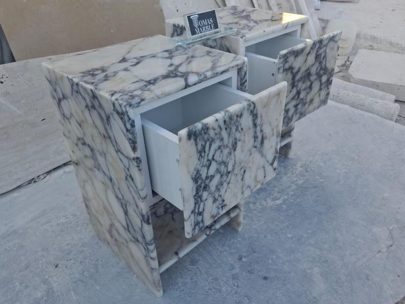 Puede incluir: Dos mesitas de noche de m&aacute;rmol con cajones abiertos. Las mesitas son blancas con vetas grises. Los cajones son blancos. El texto "OMAS MARBLE" es visible en un peque&ntilde;o letrero encima.