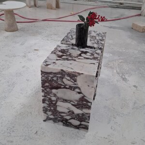 May include: A rectangular block table made of white and deep purple veined marble. A black cylindrical vase sits on the top surface, containing a small branch with bright red berries. The table has a smooth, polished finish with sharp edges.