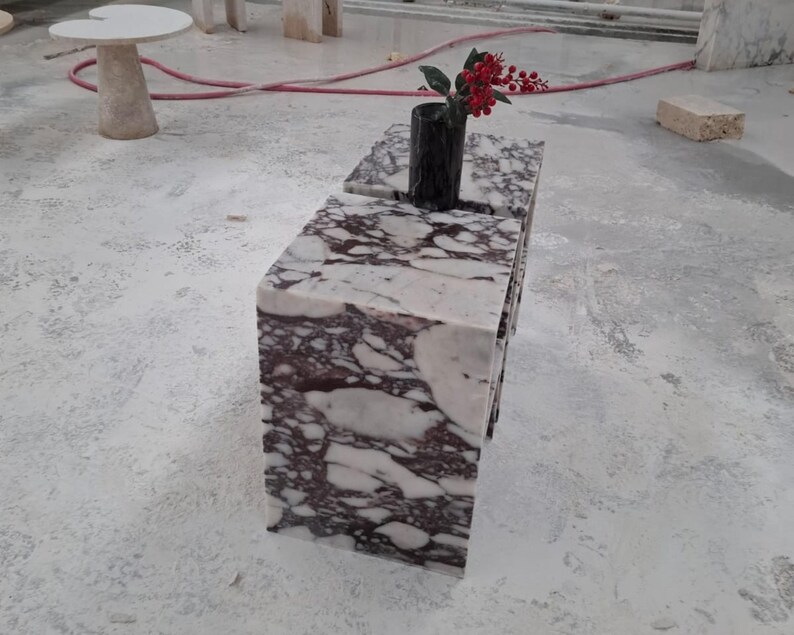 May include: A rectangular block table made of white and deep purple veined marble. A black cylindrical vase sits on the top surface, containing a small branch with bright red berries. The table has a smooth, polished finish with sharp edges.