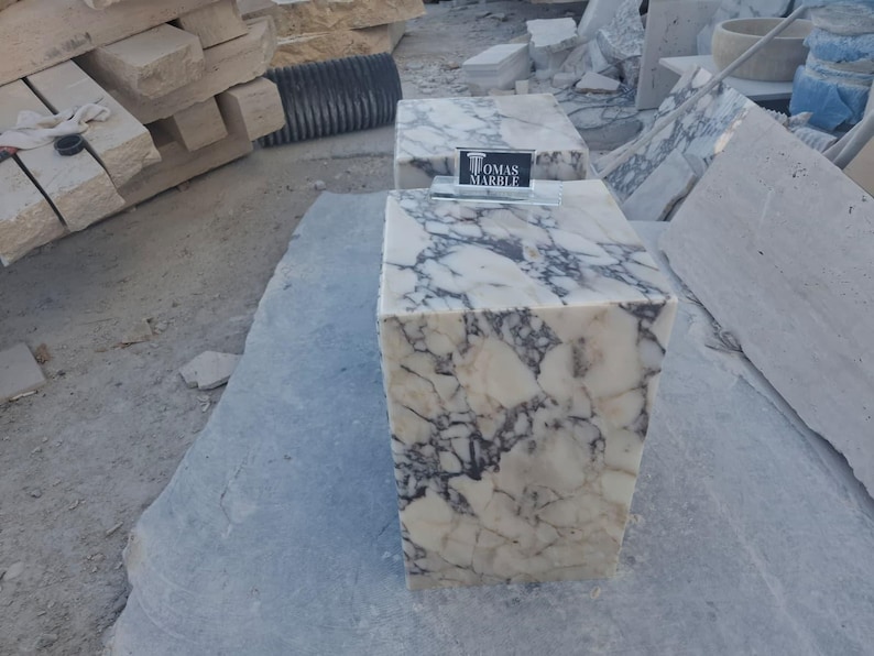Puede incluir: Dos pedestales de m&aacute;rmol con un patr&oacute;n veteado blanco y gris. Los pedestales son rectangulares y tienen el texto "TOMAS MARBLE" en un peque&ntilde;o letrero. Los pedestales est&aacute;n sobre una gran losa de piedra gris.