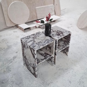 May include: Two rectangular side tables crafted from white and deep purple veined marble, each featuring a single interior shelf. A small black cylindrical marble vase containing a sprig of red berries sits between the two tables.