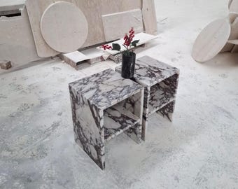 May include: Two rectangular side tables crafted from white and deep purple veined marble, each featuring a single interior shelf. A small black cylindrical marble vase containing a sprig of red berries sits between the two tables.