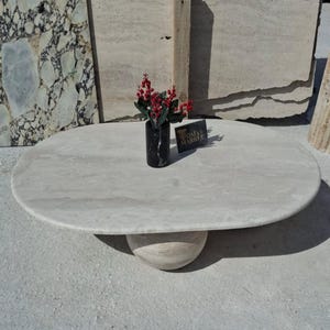 May include: A white oval marble coffee table with a round, beige marble base. The table is sitting on a light gray surface. A small vase with red berries and a black vase are on the table.