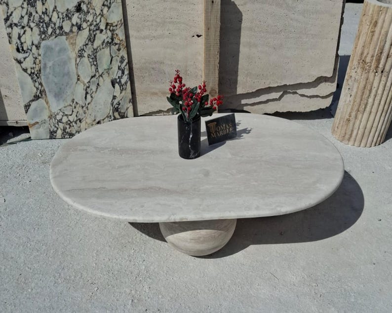 May include: A white oval marble coffee table with a round, beige marble base. The table is sitting on a light gray surface. A small vase with red berries and a black vase are on the table.