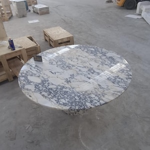 Pode incluir: Uma grande mesa redonda de mármore com uma superfície com padrões brancos e cinzentos. A mesa tem uma base sólida e está num armazém. O mármore tem um acabamento polido. O diâmetro da mesa é de aproximadamente 150 cm.