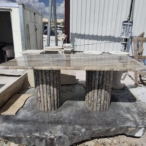 May include: A rectangular stone table with a polished, light brown, veined surface. The table is supported by two cylindrical, vertically fluted stone pedestals that feature a textured, grey and brown mottled finish throughout their structure.