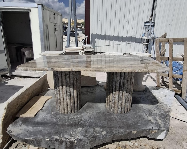 May include: A rectangular stone table with a polished, light brown, veined surface. The table is supported by two cylindrical, vertically fluted stone pedestals that feature a textured, grey and brown mottled finish throughout their structure.