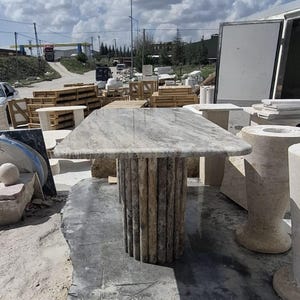 May include: A rectangular stone table featuring a polished, light grey and beige marbled top with a distinct veined pattern. The table is supported by a cylindrical base constructed from multiple vertical, textured stone columns in a neutral tone.