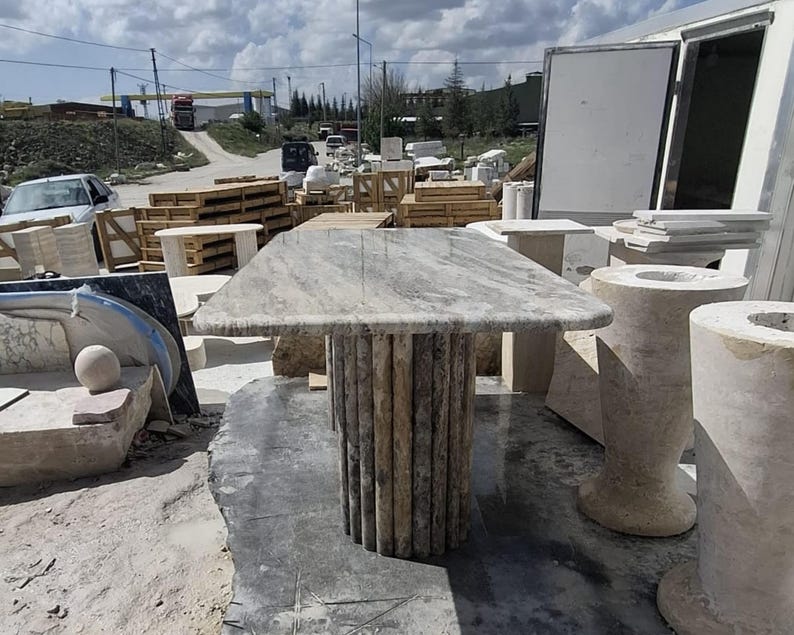 May include: A rectangular stone table featuring a polished, light grey and beige marbled top with a distinct veined pattern. The table is supported by a cylindrical base constructed from multiple vertical, textured stone columns in a neutral tone.
