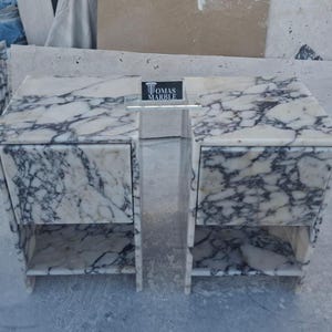 Puede incluir: Una mesita de noche de m&aacute;rmol con dos cajones y un estante abierto. La mesita es blanca con vetas grises. El nombre de la marca "TOMAS MARBLE" se muestra en un peque&ntilde;o letrero en el centro.