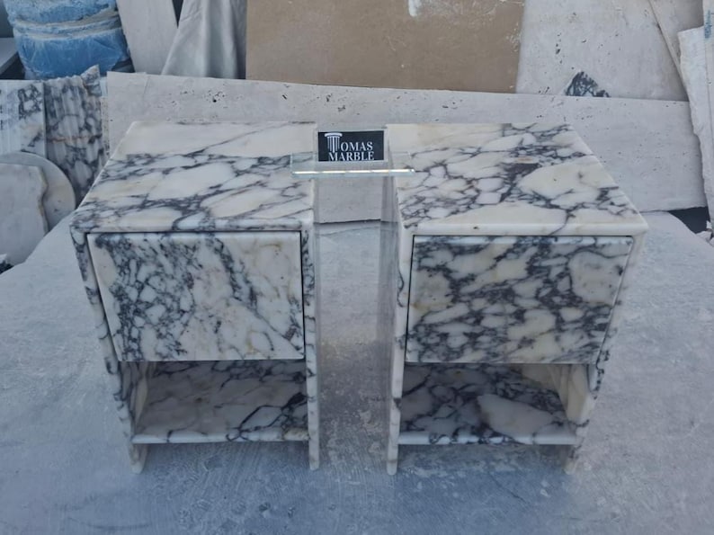 Puede incluir: Una mesita de noche de m&aacute;rmol con dos cajones y un estante abierto. La mesita es blanca con vetas grises. El nombre de la marca "TOMAS MARBLE" se muestra en un peque&ntilde;o letrero en el centro.