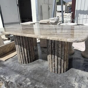 May include: A rectangular dining table featuring a polished beige and brown marble top with natural veining. The table is supported by two cylindrical bases, each constructed from a series of vertical, fluted stone columns in a matching marble.
