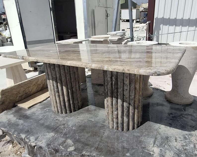 May include: A rectangular dining table featuring a polished beige and brown marble top with natural veining. The table is supported by two cylindrical bases, each constructed from a series of vertical, fluted stone columns in a matching marble.