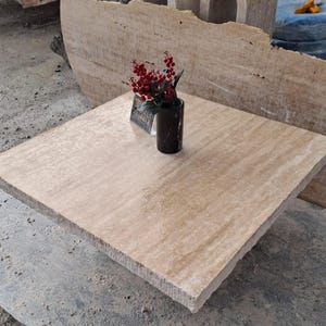 May include: A square, light brown travertine table top with a vase of red berries and a black vase on top. The table top is smooth and has a slight sheen.