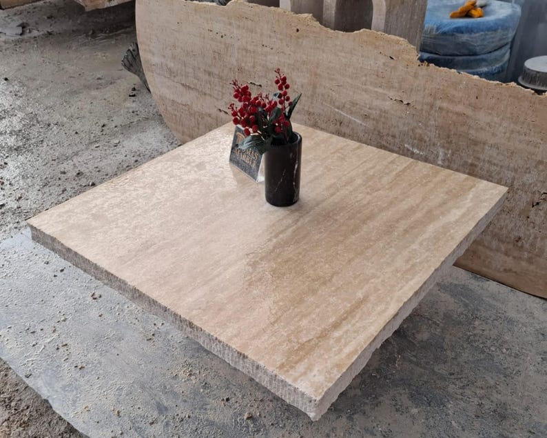 May include: A square, light brown travertine table top with a vase of red berries and a black vase on top. The table top is smooth and has a slight sheen.