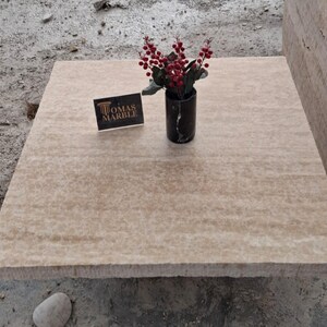 May include: A close-up of a beige travertine tile with a subtle veined pattern. The tile is sitting on a rough, gray surface. A small bouquet of red berries is in a black vase on the tile. A business card with the text "Thomas Marble" is also on the tile.