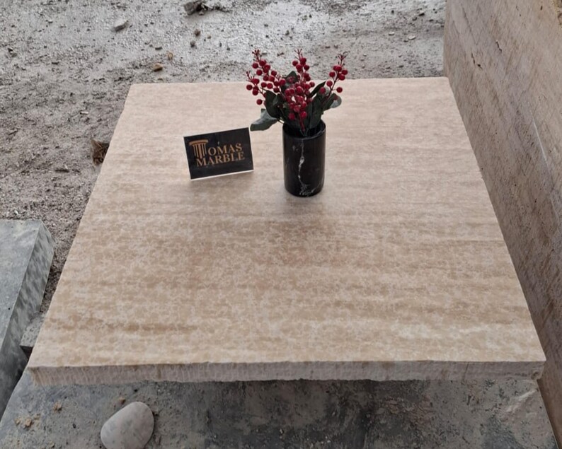 May include: A close-up of a beige travertine tile with a subtle veined pattern. The tile is sitting on a rough, gray surface. A small bouquet of red berries is in a black vase on the tile. A business card with the text "Thomas Marble" is also on the tile.