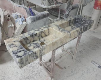 May include: A marble sink with a rectangular basin and a smaller, adjacent basin. The sink features a white and gray veined pattern. It is supported by a wooden stand in a workshop setting. The sink's dimensions are approximately 60 inches long.
