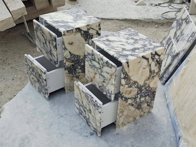 May include: Two matching bedside tables crafted from polished stone with a distinct black, white, and cream marbled pattern. Each unit features a rectangular frame and two open drawers with white interior sides, showcasing a natural stone texture.