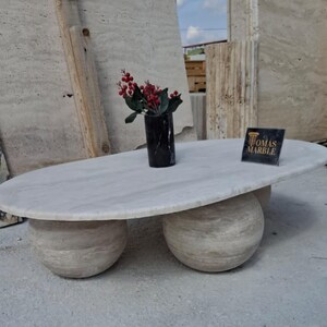 May include: A white oval-shaped coffee table with three round legs made of the same stone. The table is sitting on a concrete floor. A small bouquet of red berries is in a black vase on the table. A sign with the text "Tomas Marble" is on the table.