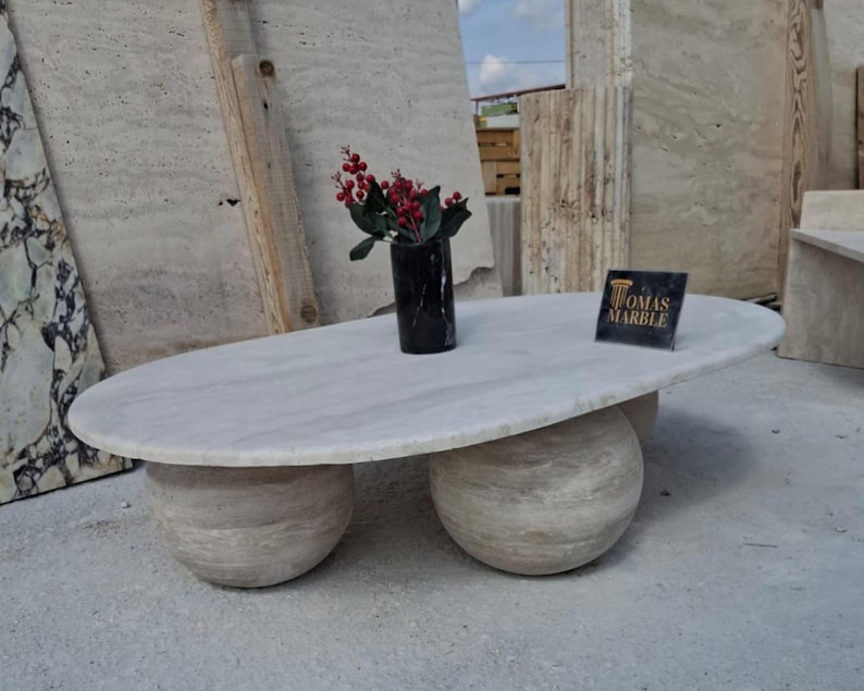 May include: A white oval-shaped coffee table with three round legs made of the same stone. The table is sitting on a concrete floor. A small bouquet of red berries is in a black vase on the table. A sign with the text "Tomas Marble" is on the table.