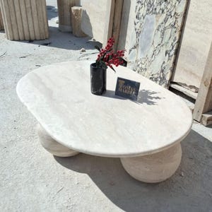 May include: A white oval-shaped coffee table with a smooth, polished surface. The table has three round legs made of the same white stone. The table is sitting on a concrete surface. A small black vase with red berries is on the table, along with a business card that reads "Tomas Marble".