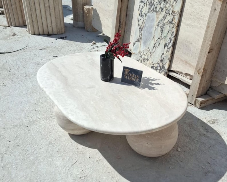 May include: A white oval-shaped coffee table with a smooth, polished surface. The table has three round legs made of the same white stone. The table is sitting on a concrete surface. A small black vase with red berries is on the table, along with a business card that reads "Tomas Marble".