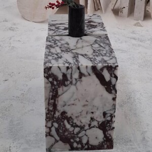 May include: A rectangular block table made of white marble with prominent dark purple and black veining. A small, cylindrical black marble vase sits on the top surface, containing a single stem with small red berries at the very top.